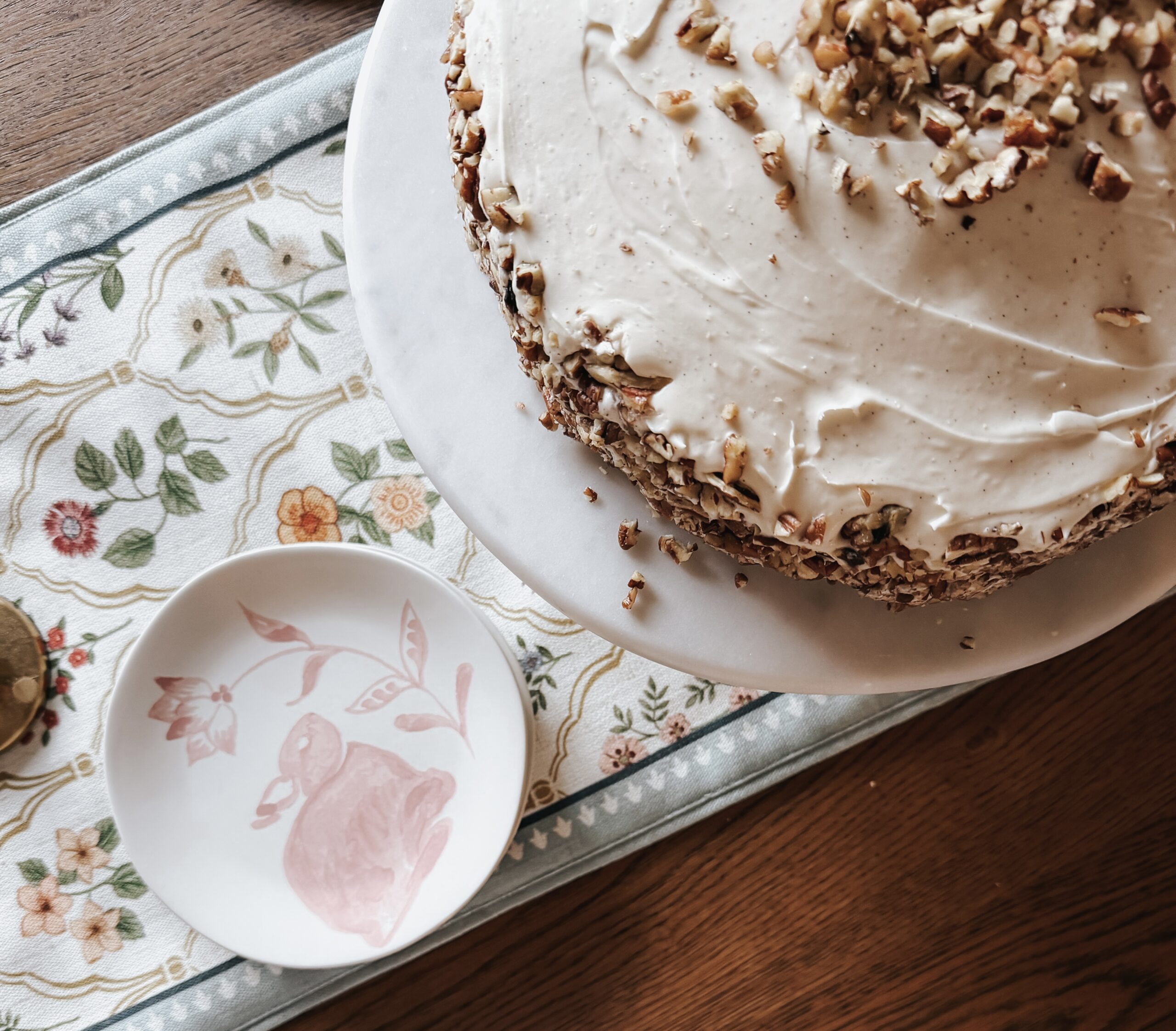 Homemade carrot cake with cream cheese frosting and salted pecans on marble cake stand with Easter table runner